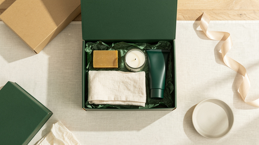 Overhead flat lay of an open deep-green gift box filled with neatly arranged soap, a candle, a hand cream tube and a fol
