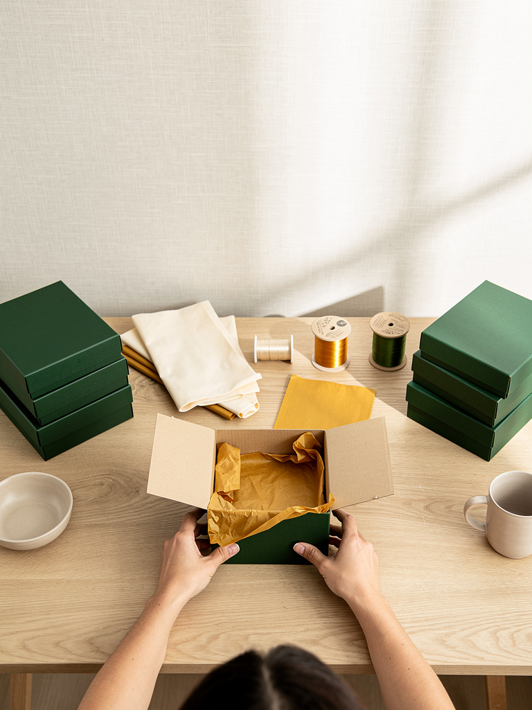 Hands seen from behind assembling a gift box at a long oak worktable lined with tidy stacks of boxes, tissue paper and r